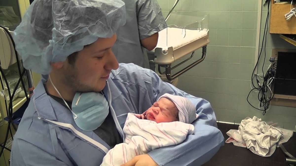 a man in gown holding a new born baby supporting his partner