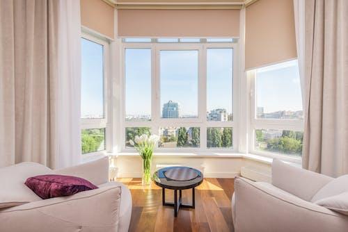 Free Interior of modern penthouse with comfortable white armchairs and round side table placed near windows overlooking city in sunny day Stock Photo